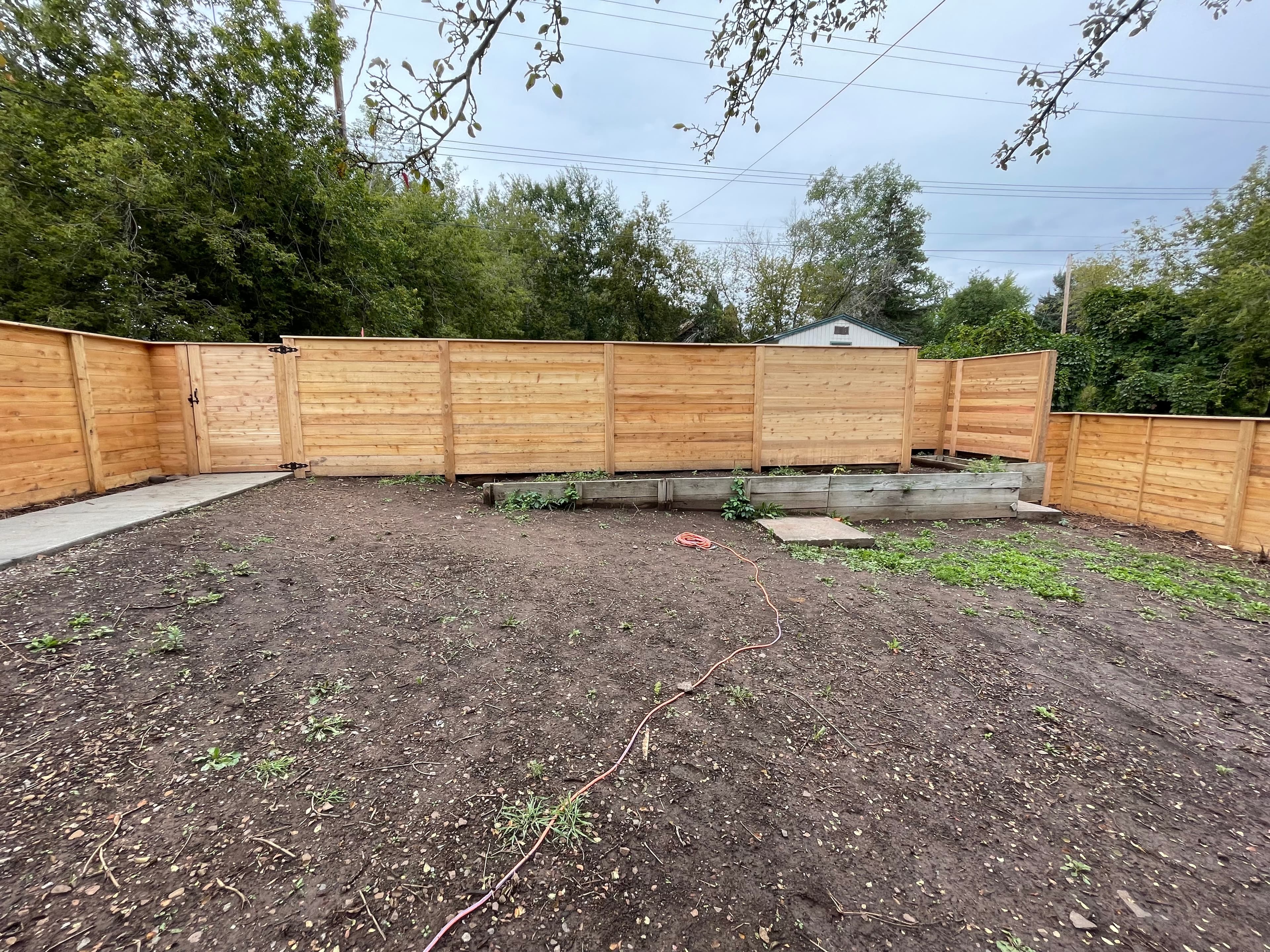 Horizontal wooden fence enclosing a dirt backyard with green trees under a cloudy sky.