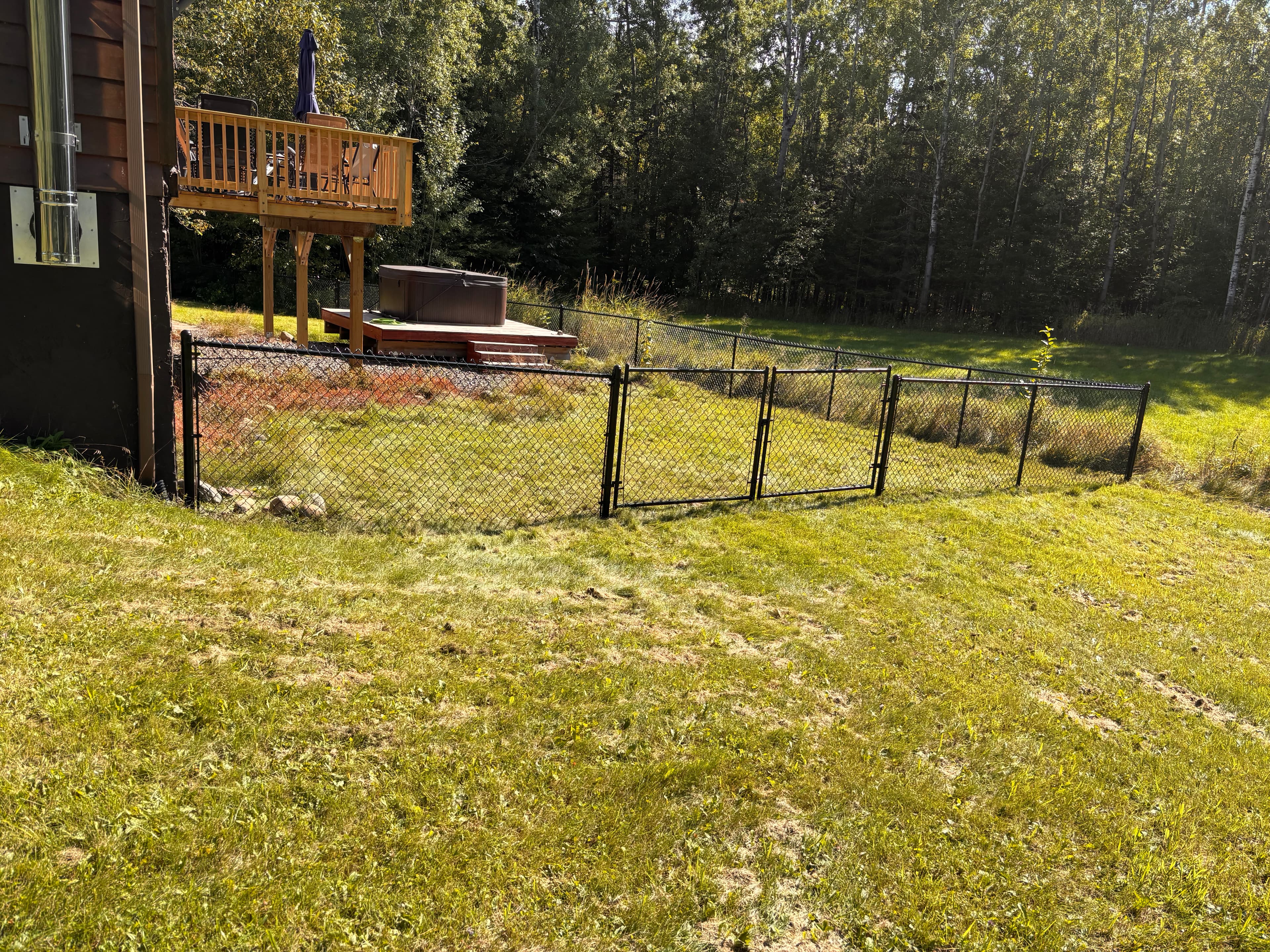 Chain-link fence in a grassy yard with a wooden deck, hot tub, and forest.