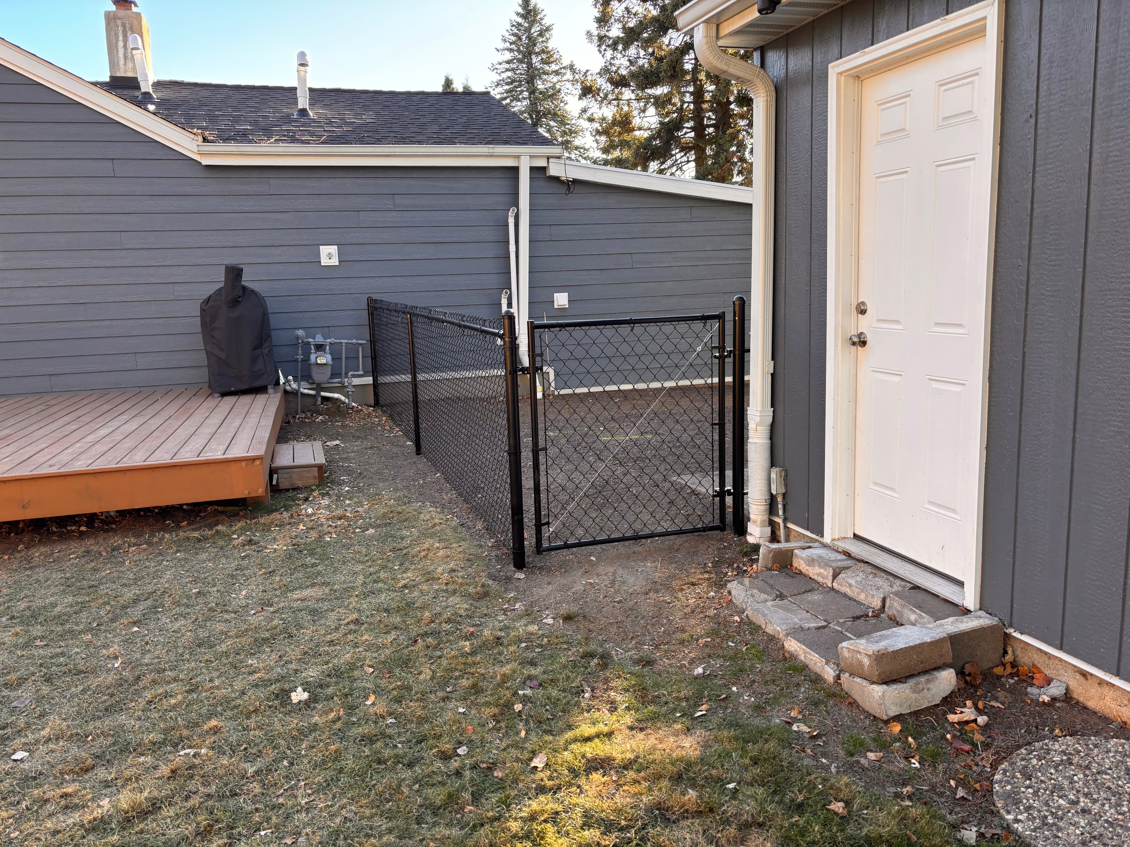 Grey house with a white door, stone steps, wooden deck, and black chain-link fence.